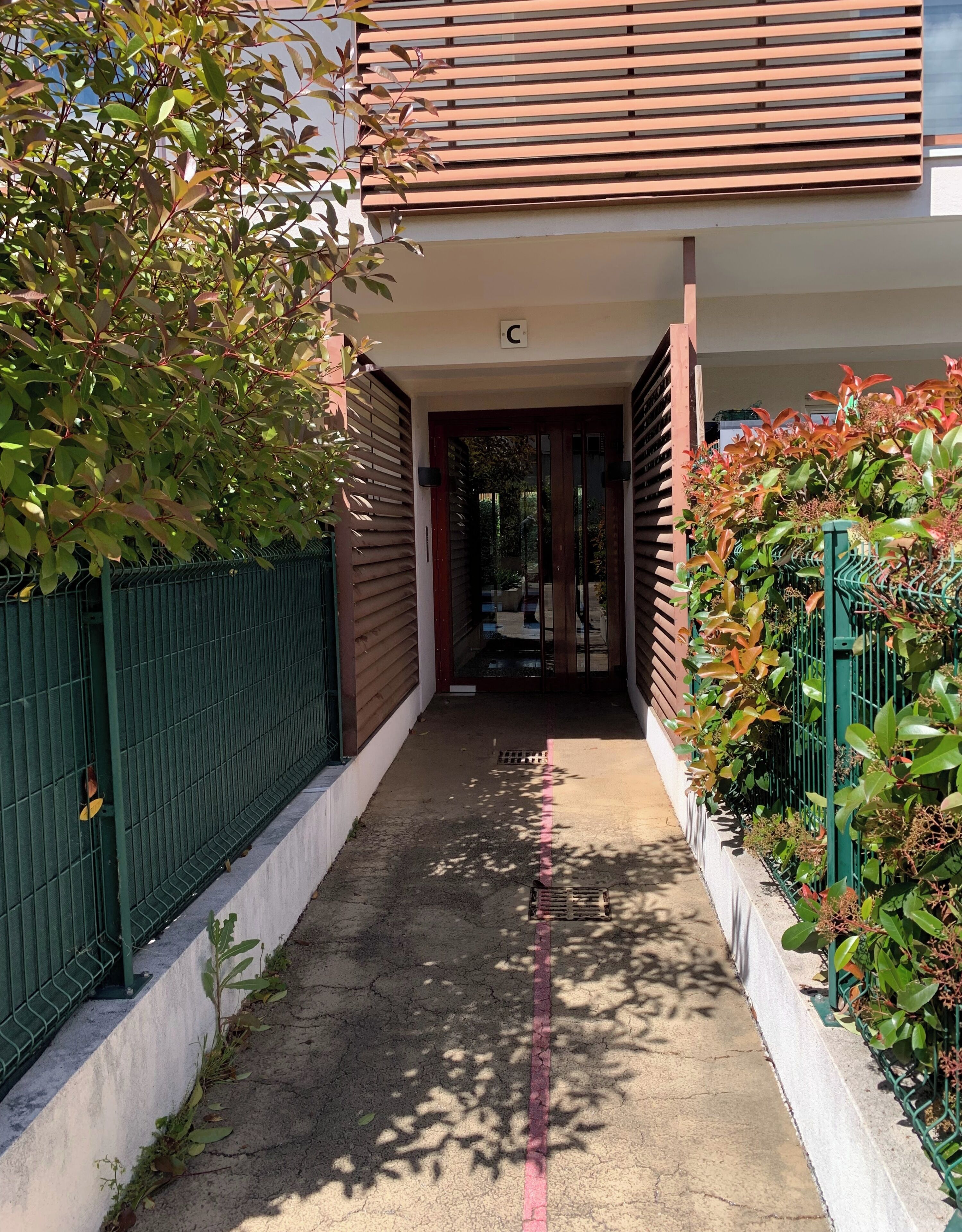 A narrow pathway leading to a building entrance with a wooden facade and a green metal fence on one side.