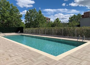 Una piscina rettangolare con un bordo in legno, una recinzione metallica e alberi sullo sfondo.