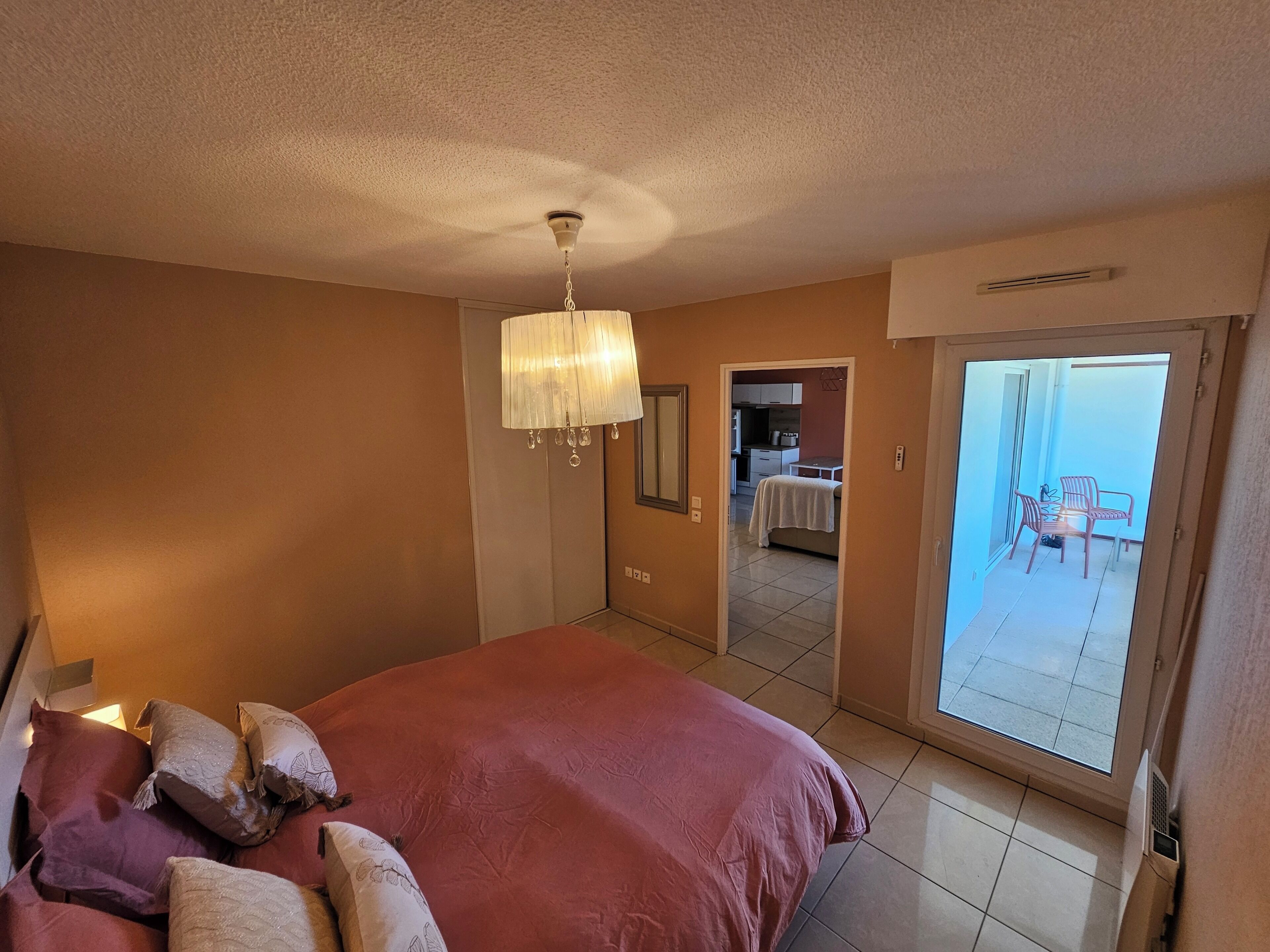 Een slaapkamer met een bed, een kroonluchter, een balkon met stoelen, en een keuken zichtbaar door een geopende deur.