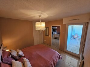Un dormitorio con cama, un candelabro, un balcón con sillas y una cocina visible a través de una puerta abierta.