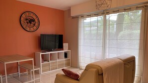 A living room with a sofa, TV, white cabinet, and a window with curtains. - Cosy apartment with pool near the center (Draguignan)