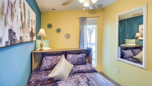Un dormitorio con cama, ventilador de techo, ventana con cortinas, mesita de noche con lámpara y espejo en la pared.