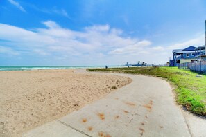 A beach with a concrete path, a grassy area, and a building with balconies. - Corpus Christi Ocean-view Condo, Walk to the Beach (Corpus Christi)
