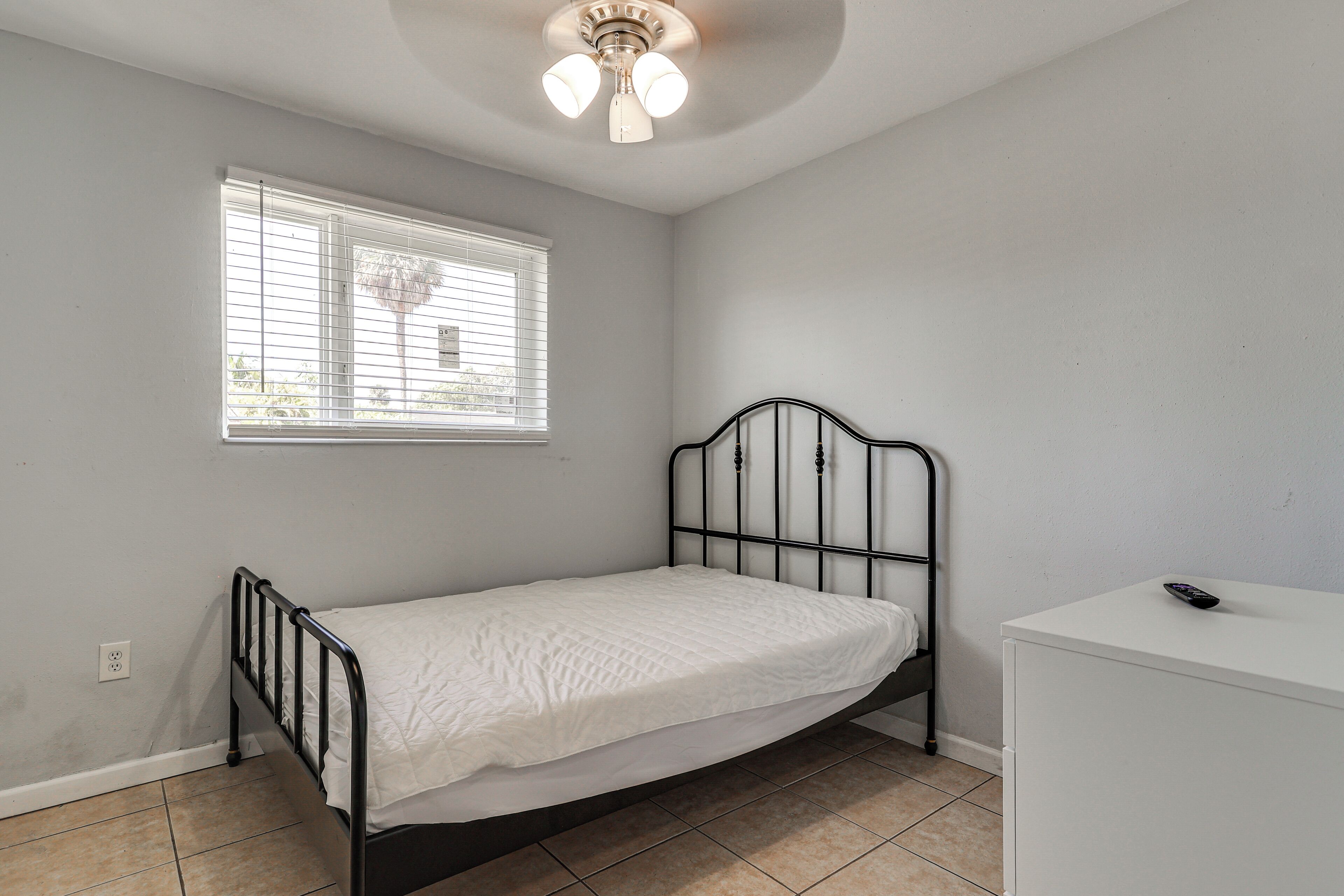 Un lit simple avec un cadre en métal, une petite commode blanche, une fenêtre avec des stores et un ventilateur de plafond.