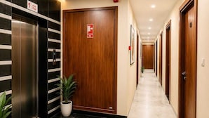 A hotel corridor with an elevator, a fire exit door, and a long hallway with doors on one side. - Hotel Lotus Palace (Lucknow)
