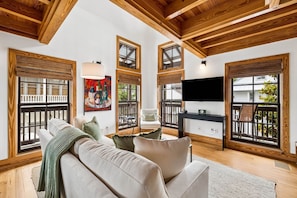 A living room with a sofa, a painting, a TV, and large windows with wooden frames.