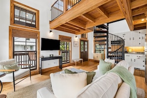 A spacious living area with a high ceiling, wooden beams, a staircase, and a kitchen with white cabinets.