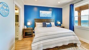 A bedroom with a bed, a nightstand, a lamp, a wall art piece, and a view of the sea through a window. - Houghton Lake Getaway: Dock, Foosball & Yard! (Houghton Lake)