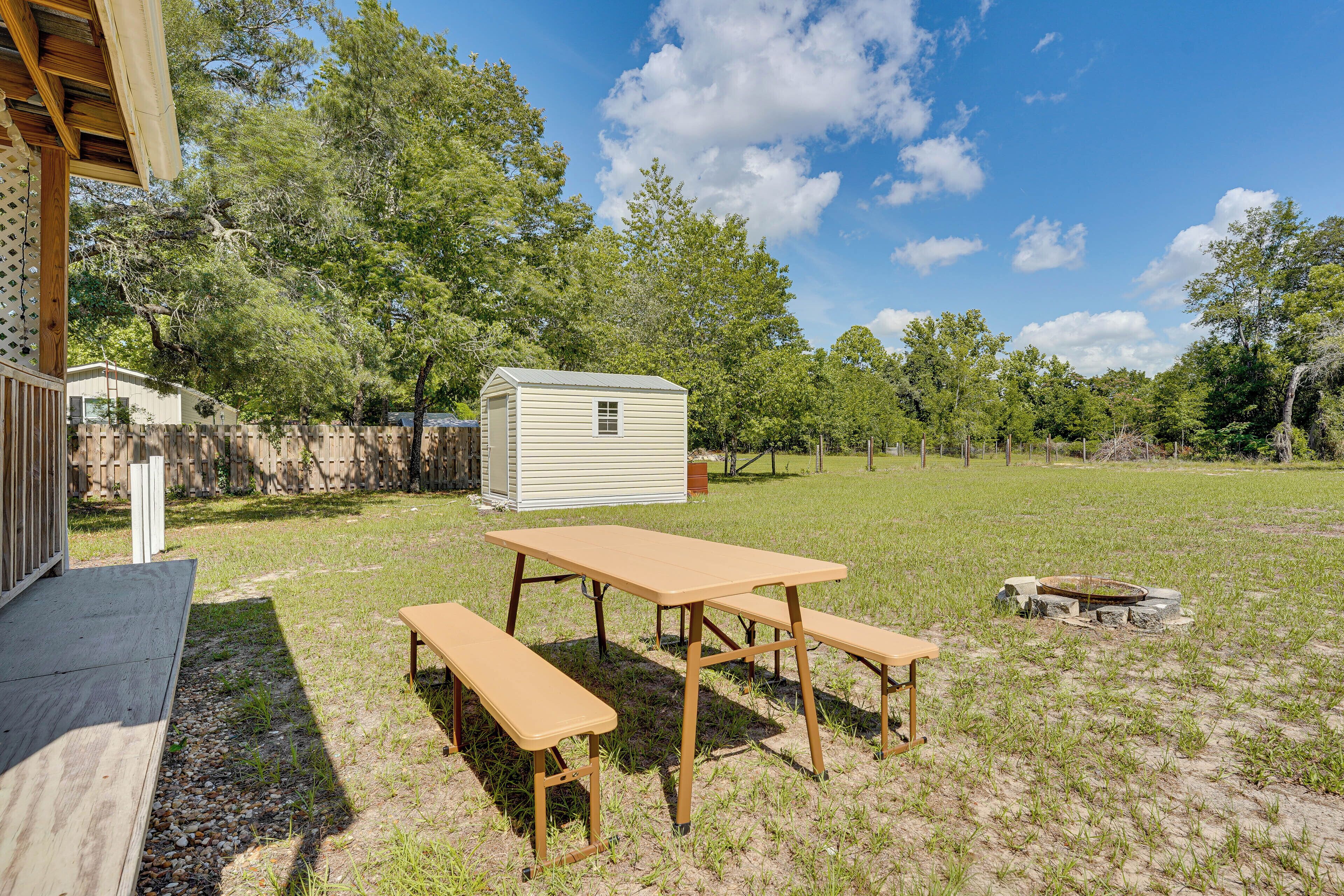Une table de pique-nique en bois, avec des bancs, se trouve dans une cour arrière verdoyante, avec des arbres et un abri de jardin en arrière-plan.