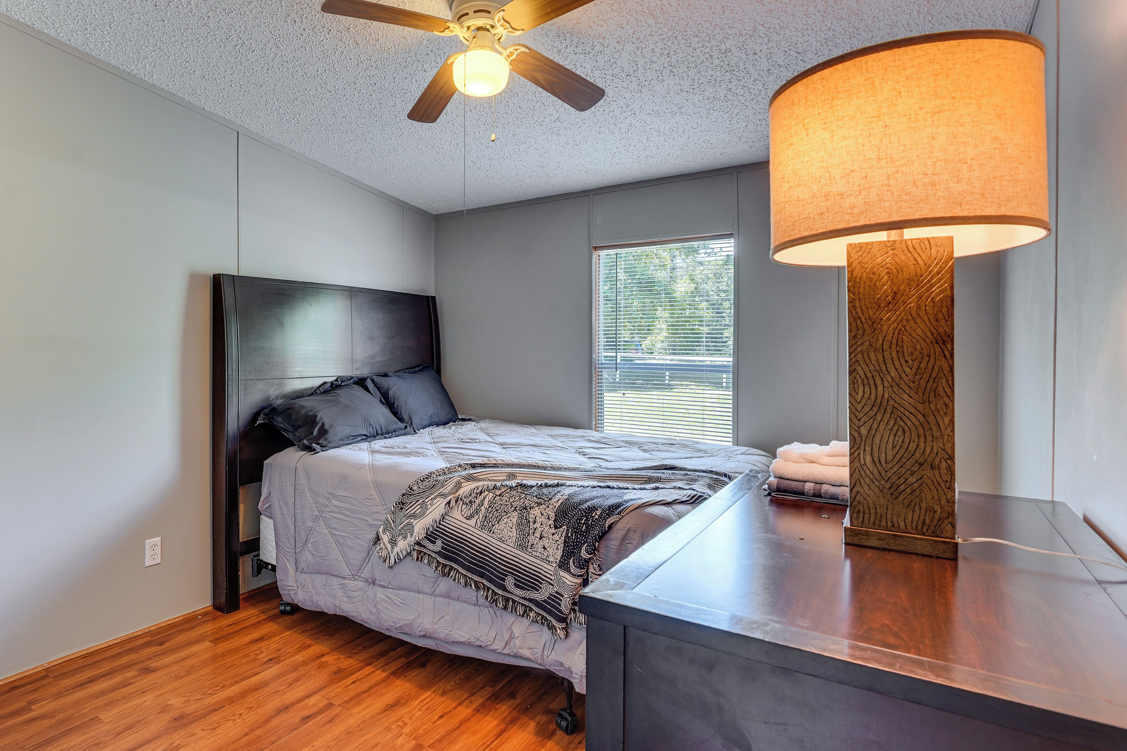 A bedroom with a bed, a ceiling fan, a window with blinds, and a bedside table with a lamp.