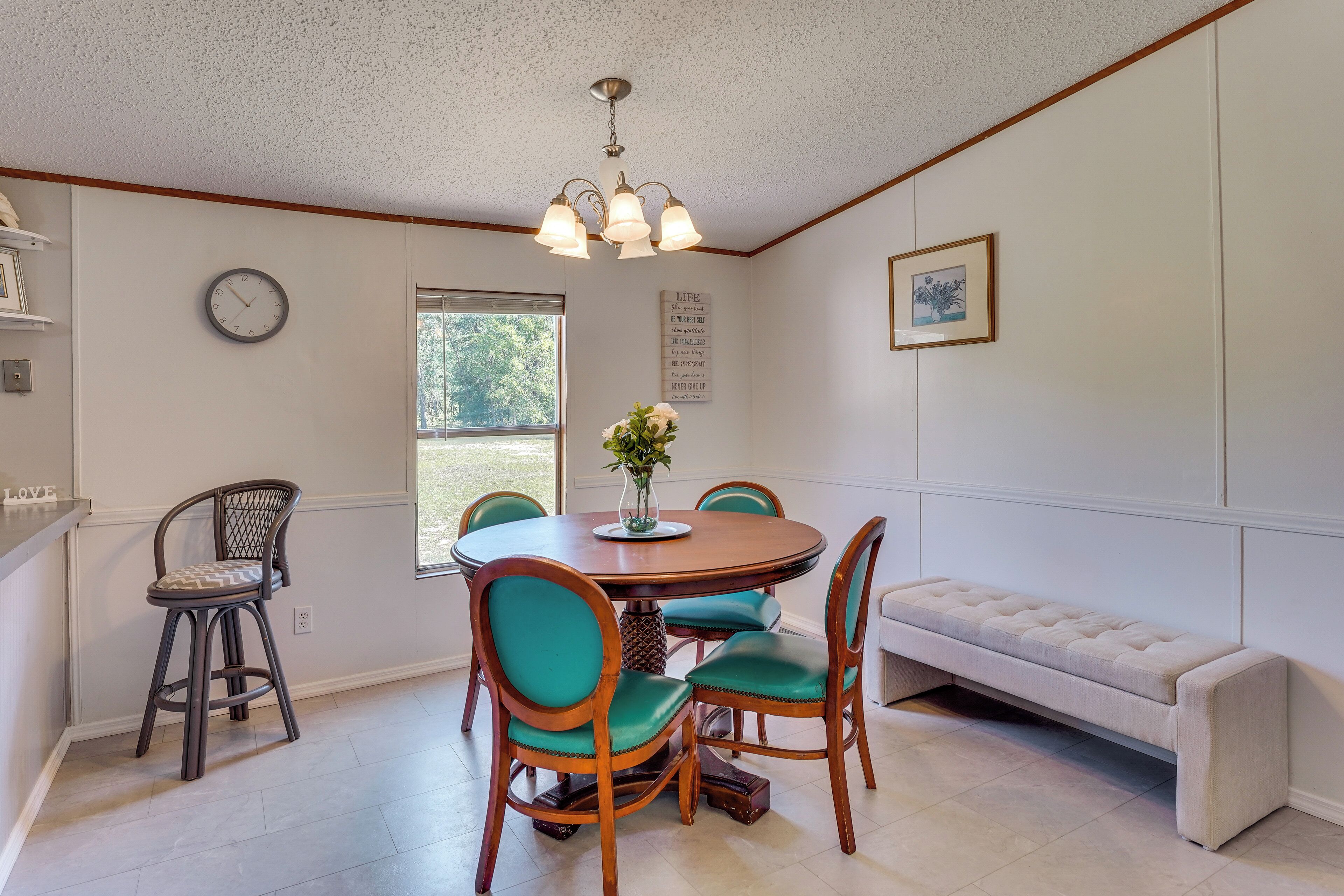A dining area with a round table, four chairs, a bench, a clock, and a window.