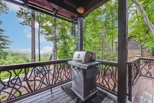 A covered outdoor area with a grill, wooden deck, and a view of a forested landscape.