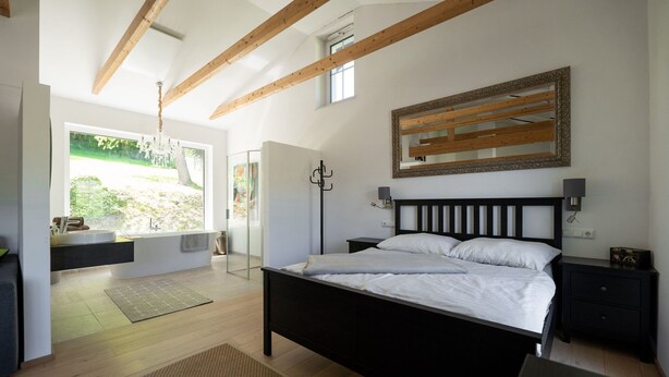 A bedroom with a bed, bedside tables, a nightstand, a mirror, and a window with a view of greenery. - WaldLoft - Southern Styria (Heimschuh)
