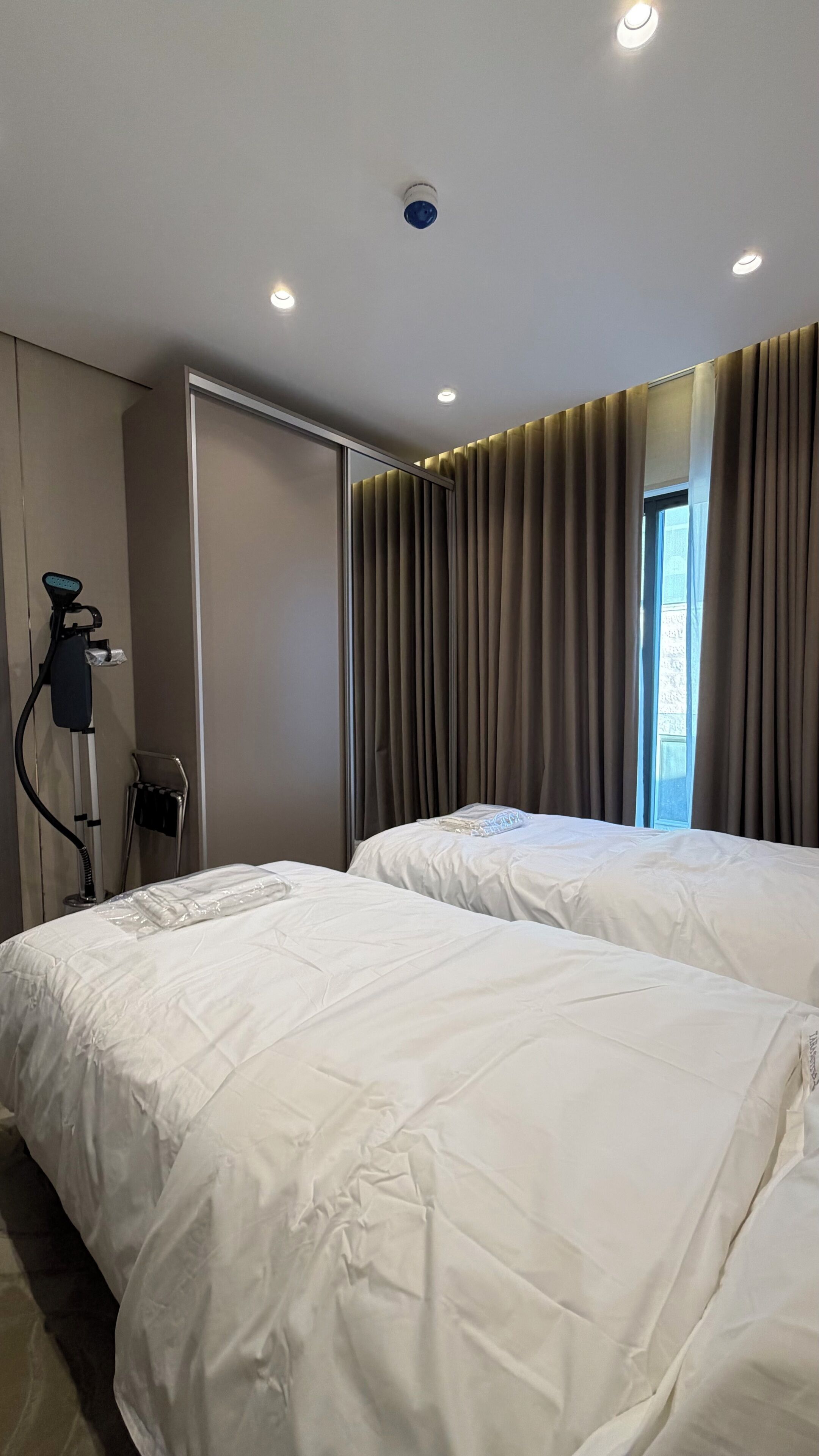 Quarto de hotel com duas camas, janela com cortinas, porta e teto com iluminação embutida.
