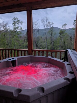 Un bain à remous éclairé d’une lumière rouge, entouré d’une terrasse en bois et offrant une vue sur une chaîne de montagnes boisées.