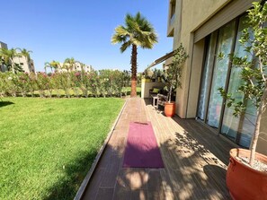 Eine Terrasse mit Holzdeck, einer violetten Yogamatte, einem Palmenbaum und Topfpflanzen.