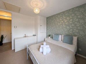 A bedroom with a bed, bedside tables, a dresser, a wall-mounted light fixture, and a door leading to another room. - Cosy ,Comfortable house with a home-from-home feel. (Hayes)