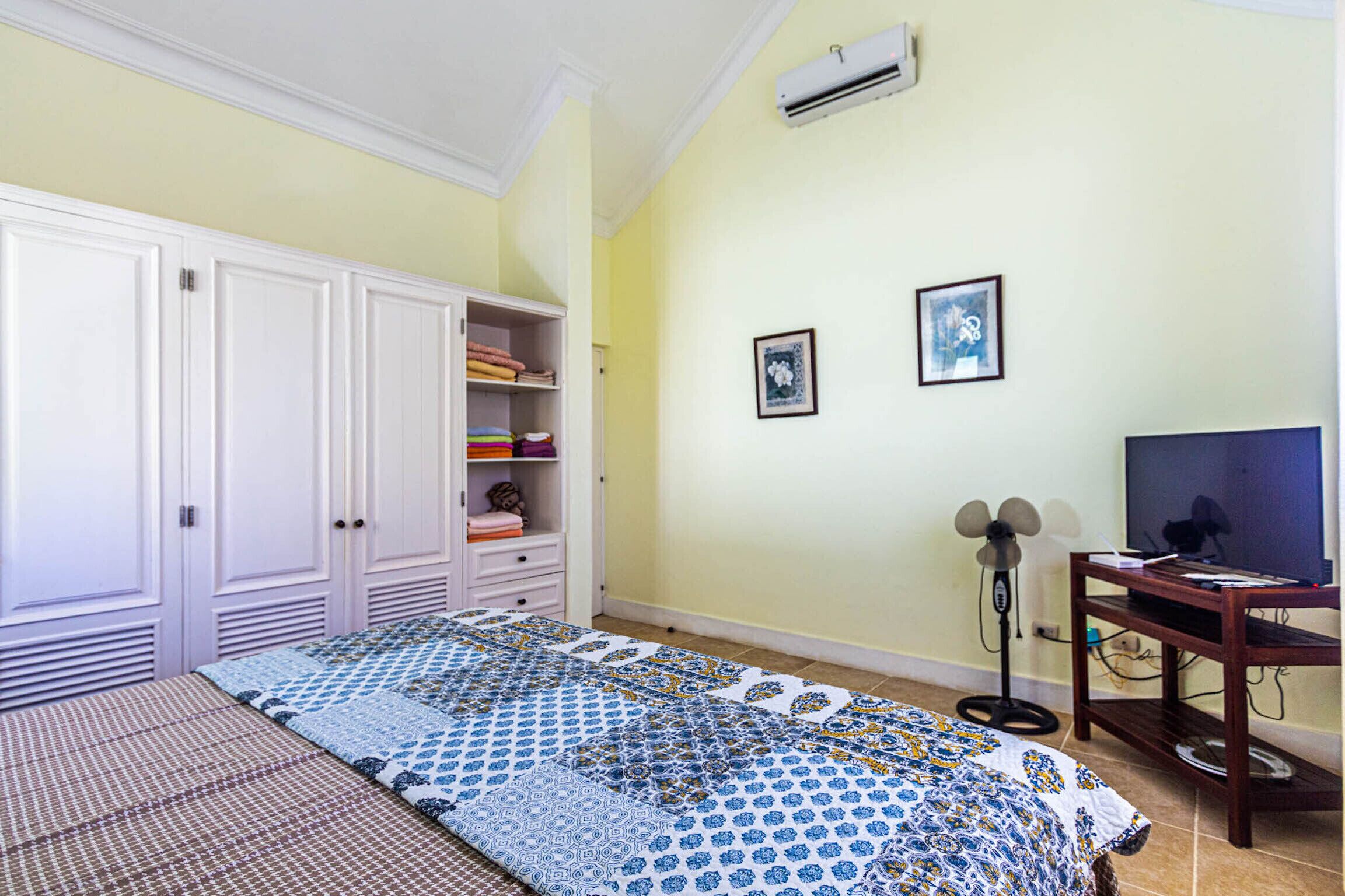 A bedroom with a bed, wardrobe, shelf with folded clothes, two framed pictures, a standing fan, and a TV on a wooden stand.