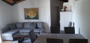 Un salón con sofá, una mesa de centro de cristal, una mesa de comedor con sillas, un cuadro de un pez y un aparador blanco con un jarrón de flores.