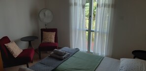 Una habitación con una cama, una silla roja, una mesita y un ventilador.