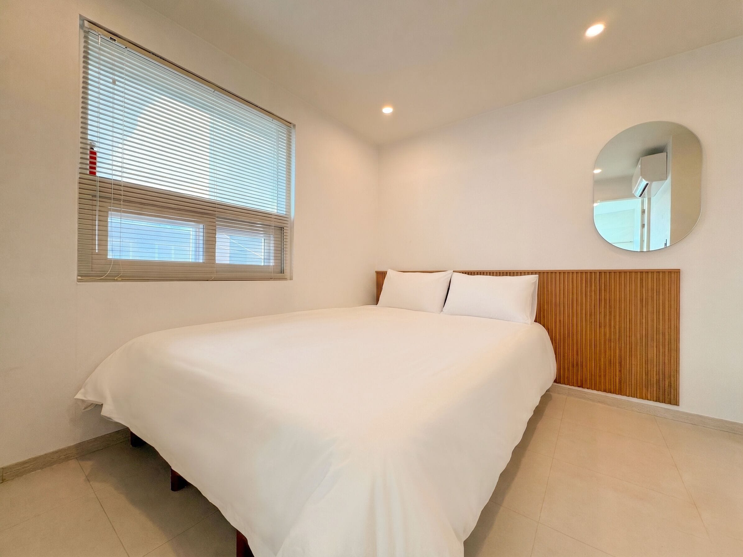 A bedroom with a bed, a window with blinds, a round mirror, and a wooden headboard.