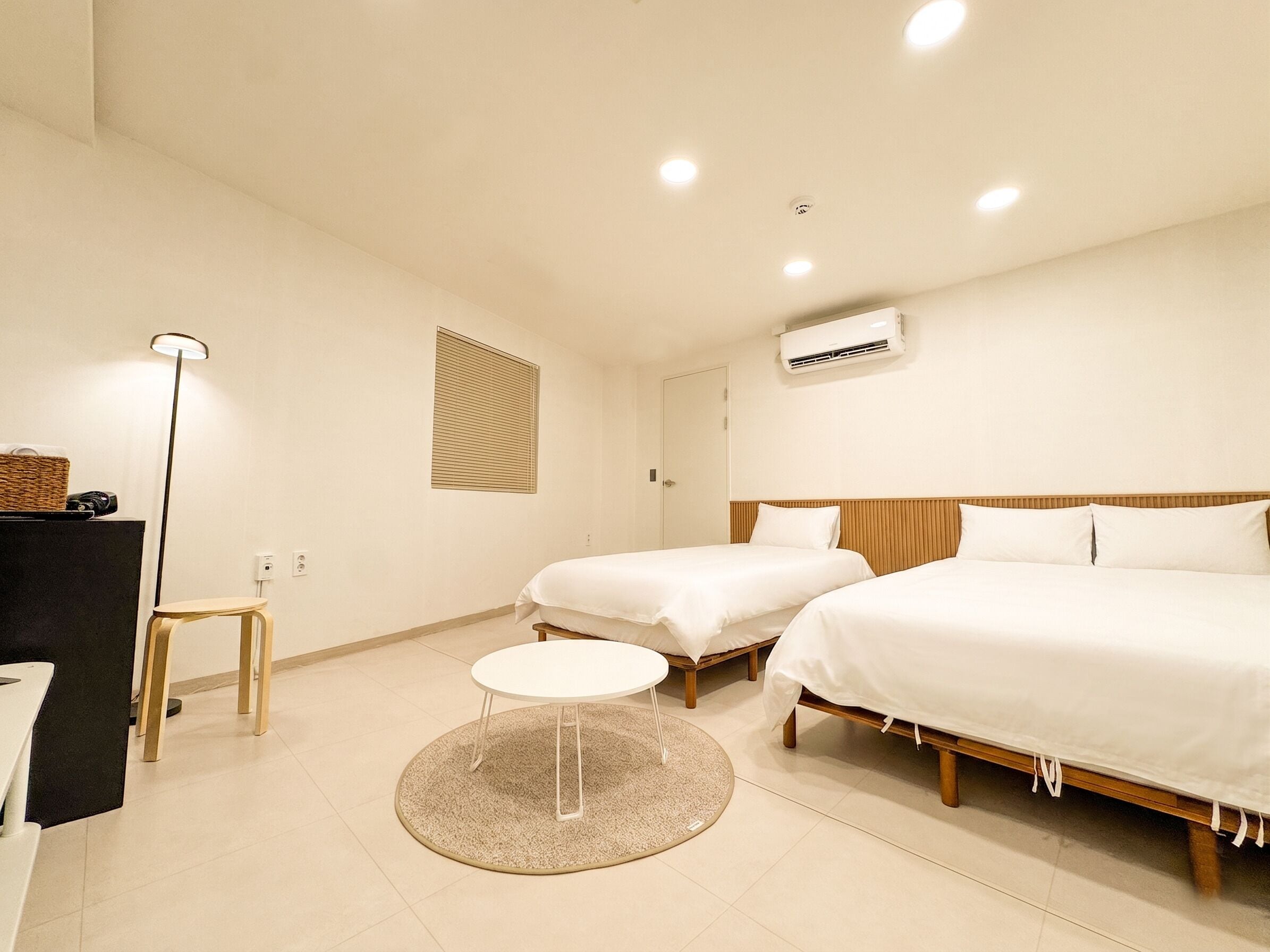 A hotel room with two beds, a small round table, a floor lamp, a desk, and a window with blinds.