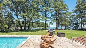Un espace aménagé au bord de la piscine, avec des fauteuils Adirondack, un barbecue et une vue sur un lac.