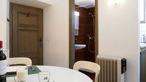 Una stanza con una porta in legno, un tavolo bianco con una bottiglia, bicchieri e un coltello, un termosifone e uno spazio doccia.
