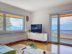 Ein Wohnzimmer mit Fernseher, Pflanze, Fernbedienung und einer Glastür, die auf einen Balkon mit Bergblick führt.