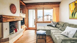 Ein gemütliches Wohnzimmer mit Kamin, Holzbalken, einer Couch und Blick auf Gebäude durch große Fenster.