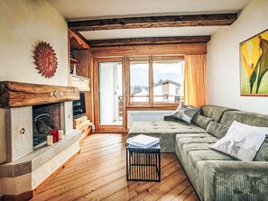 Una sala de estar acogedora con chimenea, vigas de madera, un sofá y vistas de edificios a través de amplias ventanas.