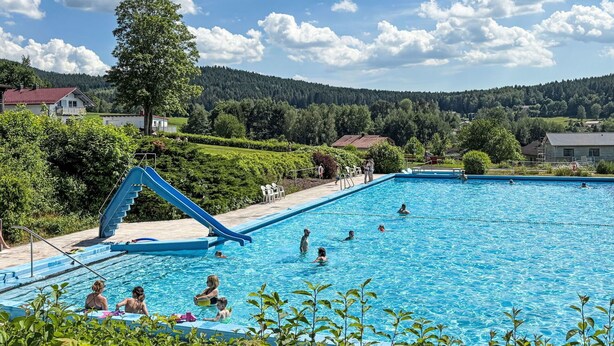 Uima-allas, jossa on liukumäki, vehreän kasvillisuuden ja talojen ympäröimänä, kirkkaan taivaan alla.