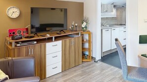 Moderni olohuone, jossa on litteänäyttöinen televisio, puinen mediakeskus ja ruokailutila. Avonaisen oven kautta näkyy keittiö.