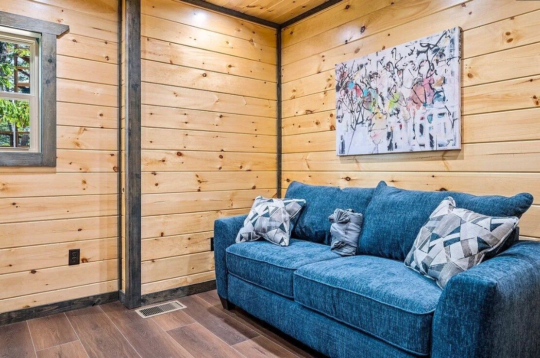 A wooden-paneled room with a blue sofa, a painting, and a window.