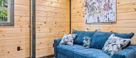 A wooden-paneled room with a blue sofa, a painting, and a window.