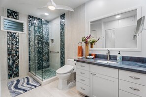 A modern bathroom with a glass shower enclosure, a white toilet, a vanity with a sink, and a mirror. - Keauhou Punahele C103 2 Bedroom Condo (Kailua-Kona)