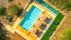 Vista aérea de una piscina rectangular con un muelle de madera, bordeada por una cerca de piedra. Hay sillas de descanso y sombrillas dispuestas a lo largo del muelle.