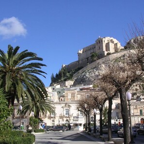 A street with buildings, palm trees, and a castle-like structure on a hill. - Wonderful private villa for 10 people with WIFI, private pool, A/C, TV, patio and pets allowed (Marina Di Ragusa)