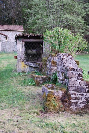 Ein rustikales Steinhaus mit einem Metallgitter, umgeben von Grünpflanzen und einem Steinbau im Hintergrund.