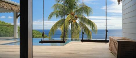 Una terraza de madera con una hamaca, una palmera y vista al océano.