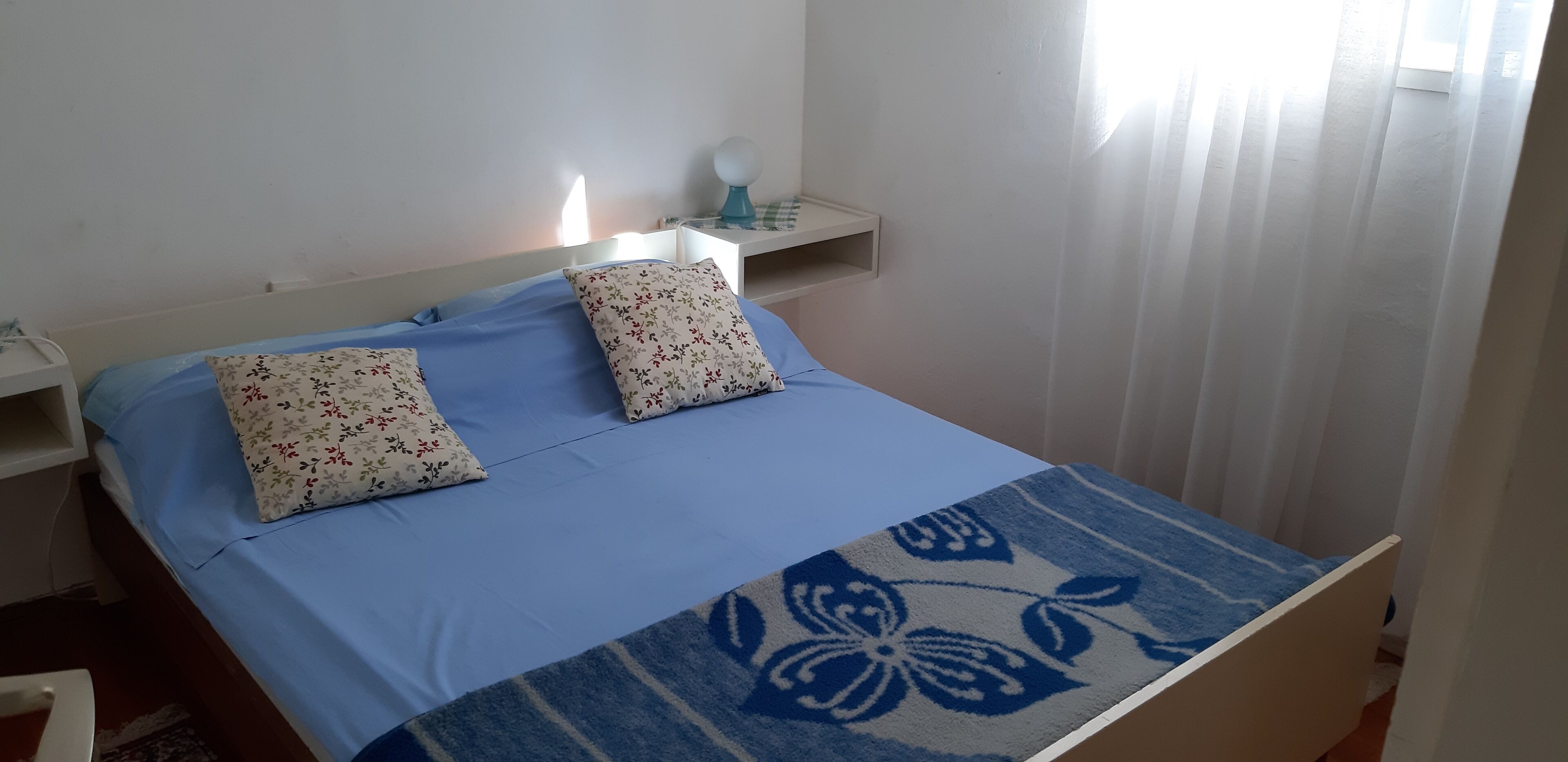 A bed with floral pillows, a blue bedspread, a bedside table with a lamp, and a window with curtains.
