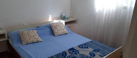 A bed with floral pillows, a blue bedspread, a bedside table with a lamp, and a window with curtains.