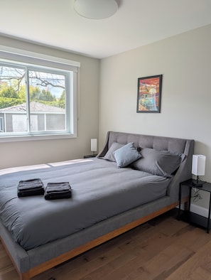 Un dormitorio con una cama grande, una mesita de noche, una lámpara de noche, una ventana con vista a árboles y una casa, y un cuadro en la pared.