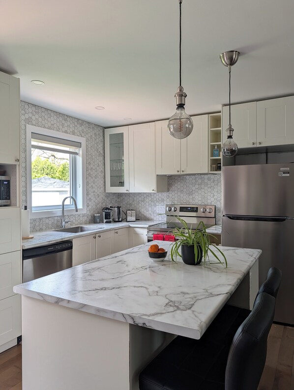 Una cocina moderna con armarios blancos, encimera de mármol, refrigerador de acero inoxidable y una lámpara colgante.