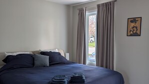 Une chambre à coucher avec un lit, des oreillers, une couverture pliée, une fenêtre avec des rideaux et un tableau encadré accroché au mur.