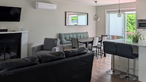 Un salon moderne avec un canapé en cuir noir, un coin repas avec une table et des chaises, un espace cuisine avec un bar, et un téléviseur fixé au mur.
