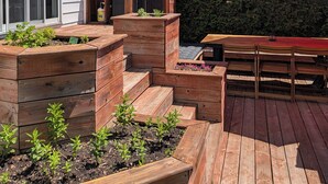 Une terrasse en bois avec des jardinières surélevées, un barbecue et un parasol rouge.