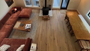 Uma sala de estar acolhedora com um sofá, mesa de centro e lareira. Grandes janelas proporcionam vista para uma floresta.
