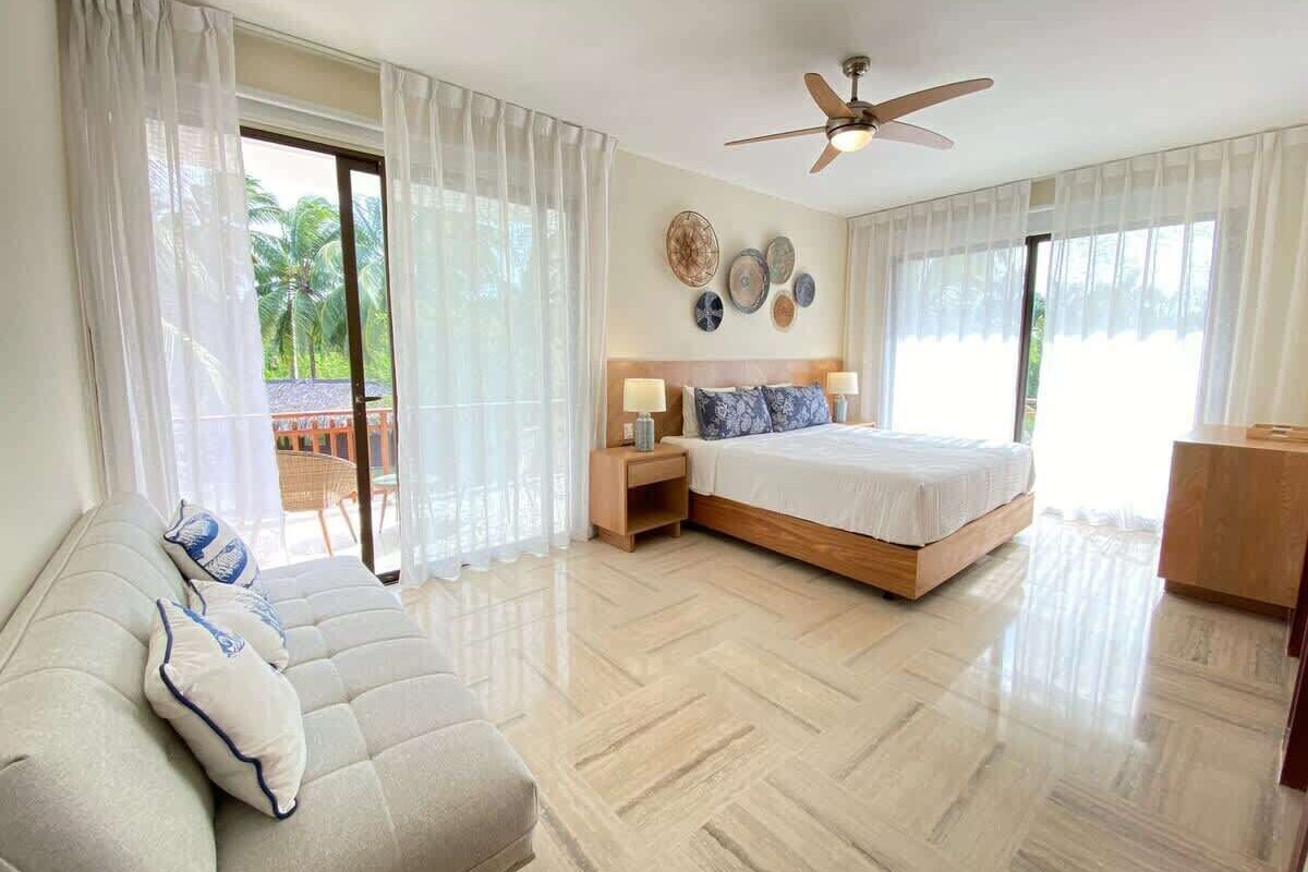 Ein Hotelzimmer mit Bett, Sofa, Deckenventilator und Blick auf Palmen.