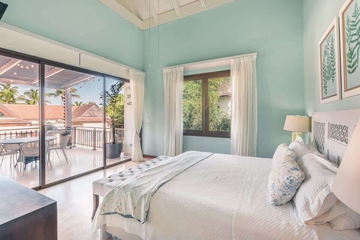 Ein Schlafzimmer mit einem großen Bett, einer Glasschiebetür zu einem Balkon, einem Fenster mit Vorhängen und gerahmten Kunstwerken an der Wand.